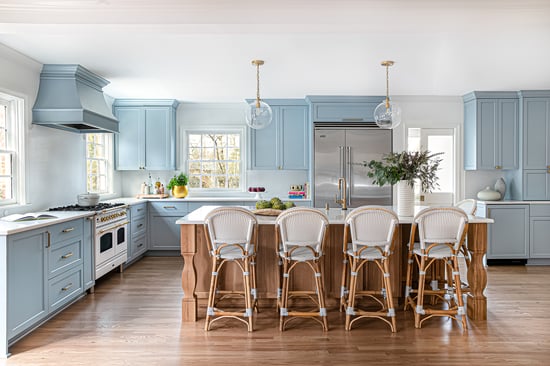 Bright coastal-inspired open-concept kitchen remodel in Myers Park, Charlotte, NC, featuring soft blue cabinetry, white countertops, a large wood island with four woven stools, a custom range hood, white-and-brass range, stainless steel refrigerator, glass pendant lights, and warm wood floors