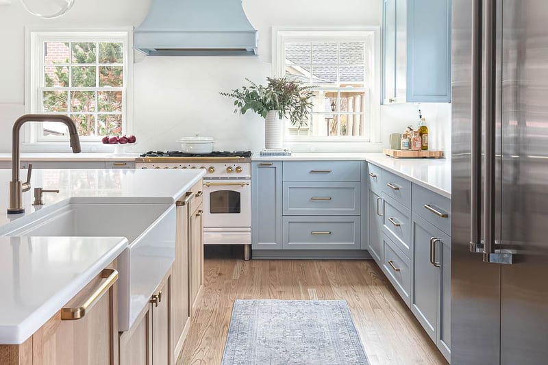 Bright coastal-style kitchen remodel in Myers Park Charlotte, NC with soft blue cabinetry, brass hardware, white countertops, and a farmhouse sink. A white-and-brass range sits beneath a custom blue range hood, while large windows fill the space with natural light