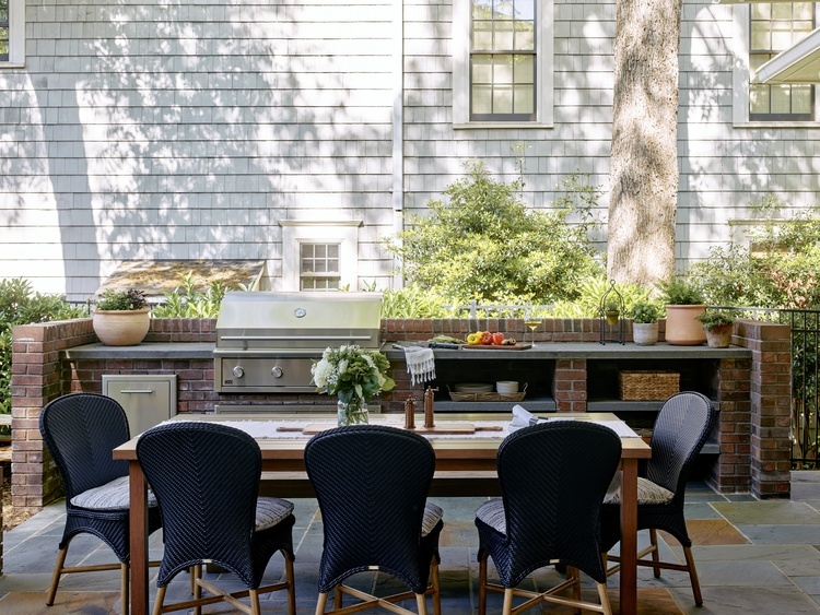 Outdoor dining patio in Dilworth, Charlotte, NC, featuring a brick outdoor kitchen with stainless steel grill surrounded by Noble Bluestone countertops, Pennsylvania bluestone flooring, landscaped surroundings, and a wood dining table with woven chairs.